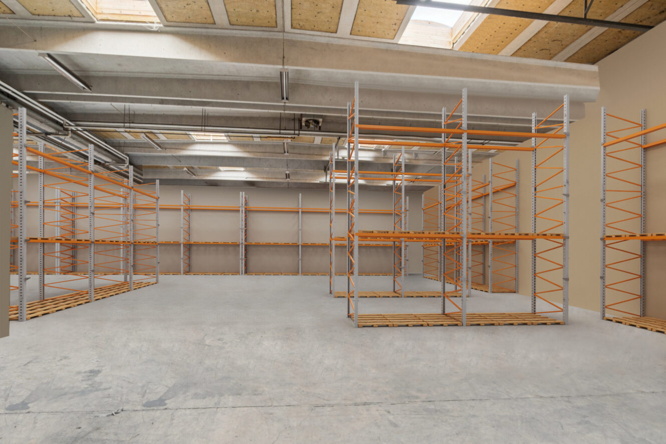 Tomt lagerinteriør med betongulv, højt til loftet og flere metalreoler med orange støtter. Naturligt lys kommer ind gennem ovenlysvinduer i taget.