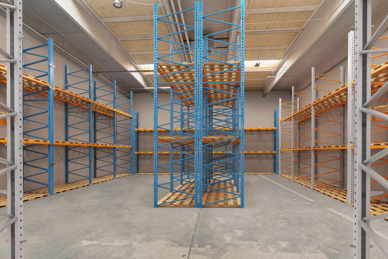 Tom lagerbygning med metalreoler i blå, orange og hvid, som er placeret rundt omkring i lokalet under et loft med ovenlys og synlige bjælker.