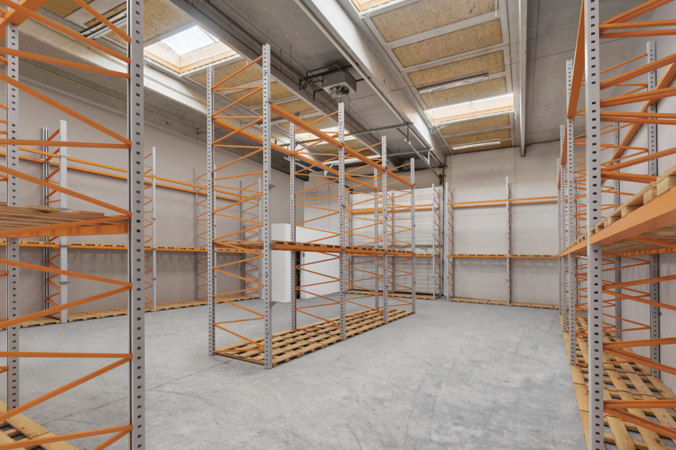 Tomt lager med metalreoler og træpaller på et betongulv, under et loft med ovenlys og synlige rør.