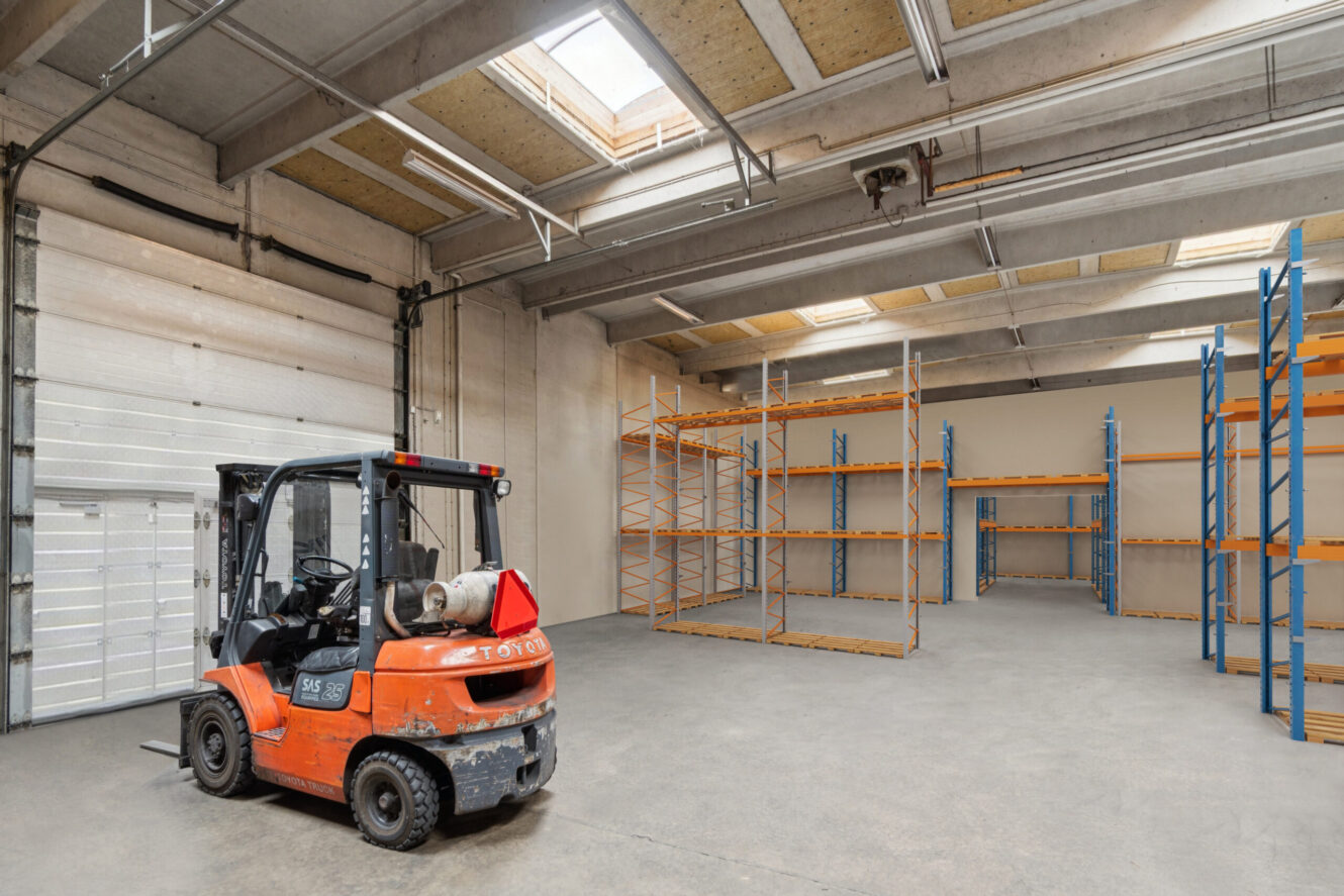 En gaffeltruck er parkeret på gulvet i en rummelig lagerhal med tomme metalreoler og store ovenlysvinduer.