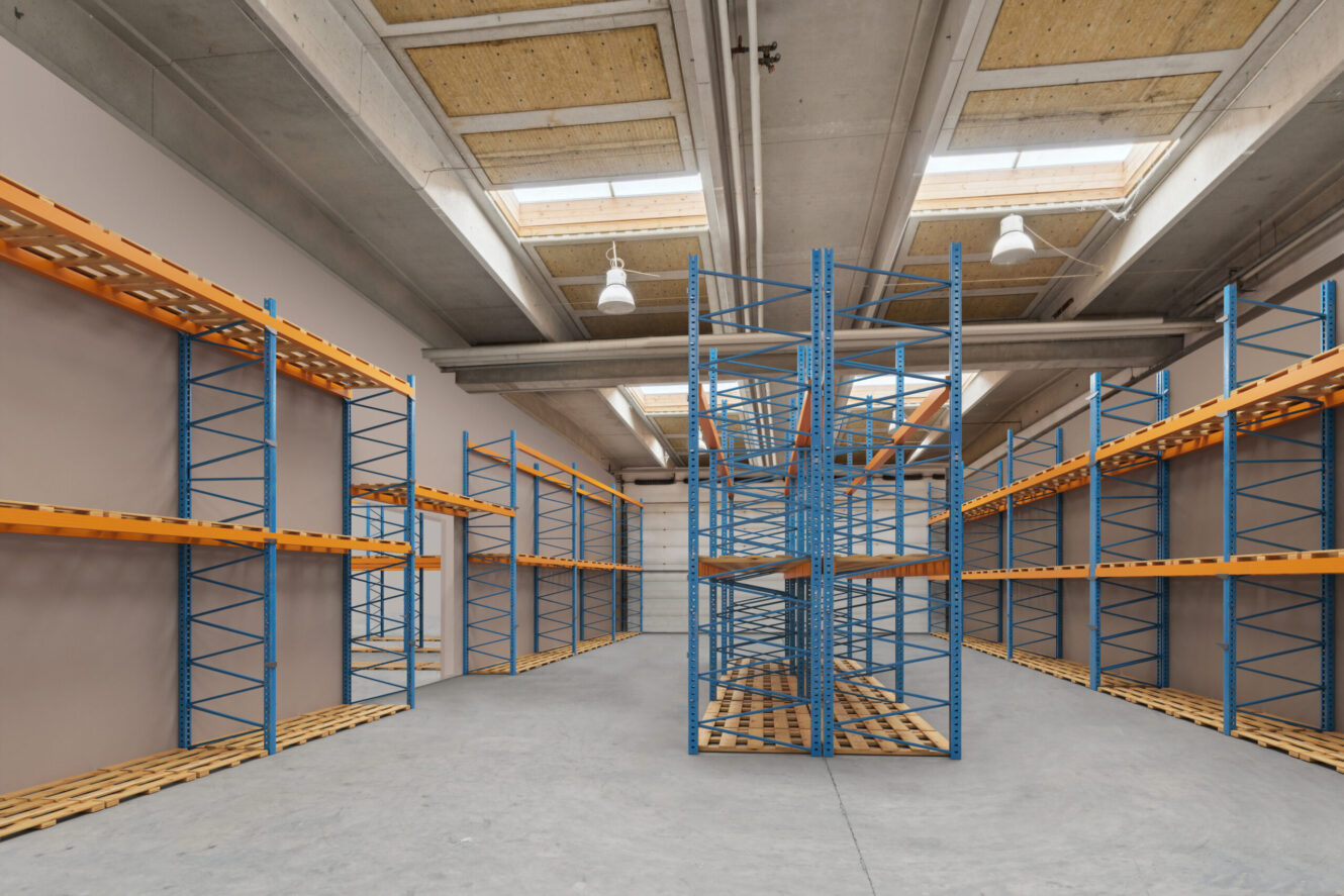 Tomme lagerreoler i metal med orange bjælker og blå rammer står i rækker i et stort industriområde med betongulv og ovenlys.