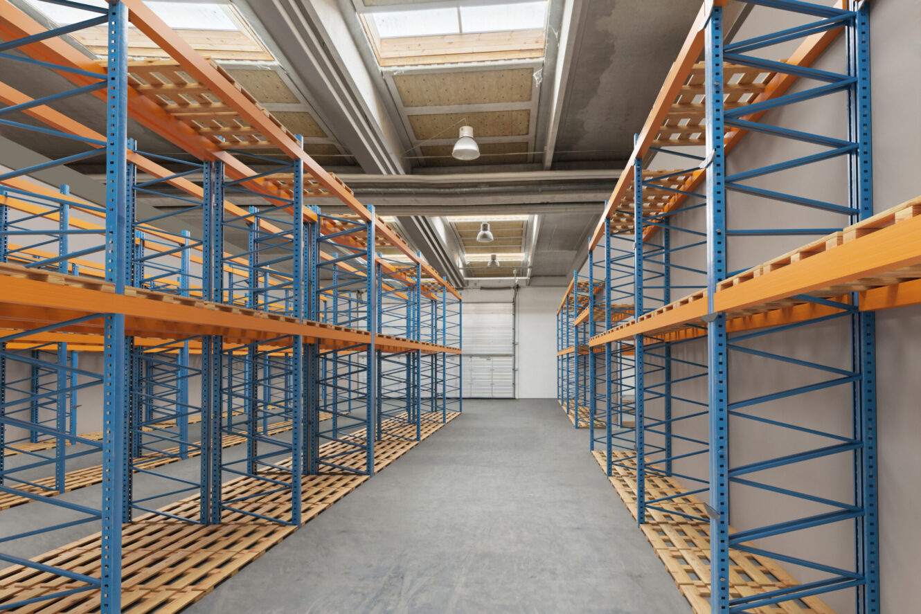 Tomt industrilager med blå og orange metalreoler og træpaller på gulvet, under et loft med ovenlysvinduer og synlige bjælker.