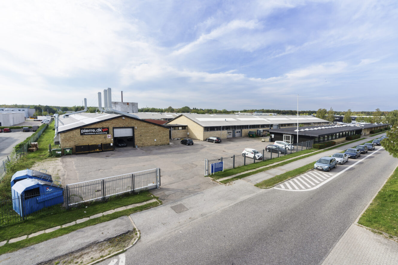 Bredt billede af en industribygning med et indhegnet parkeringsområde, mærket pierre.dk, omgivet af veje, parkerede biler og andre industrianlæg.