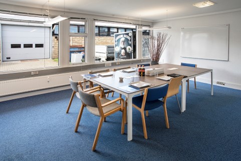 Et moderne konferencelokale med et rektangulært bord, seks stole, dokumenter, drikkevarer, et whiteboard og store vinduer, der viser et industrielt ydre.