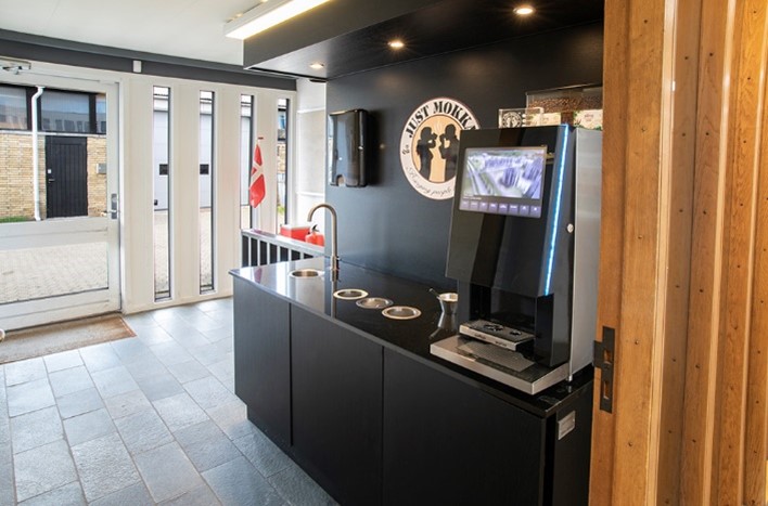 En moderne selvbetjent kaffemaskine står på en sort bordplade i et lyst, minimalistisk rum med klinkegulv og store glasdøre.