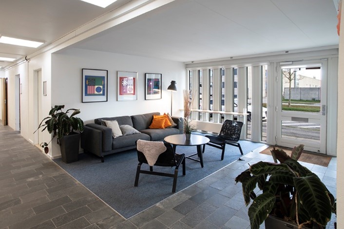 Et moderne loungeområde med en grå sofa, stole, sofaborde, indrammet vægkunst og store vinduer, der lukker naturligt lys ind.