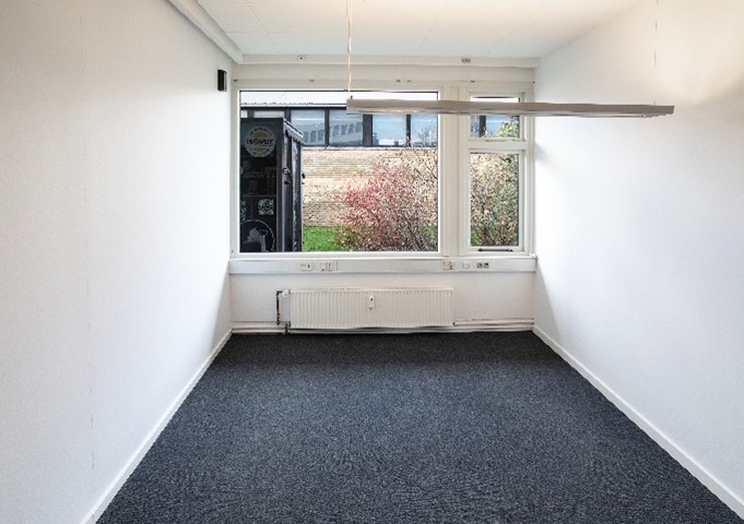 Et tomt, smalt rum med hvide vægge, et mørkt gulvtæppe, en radiator under et vindue og et hængende lysarmatur i loftet. En automat er synlig uden for vinduet.