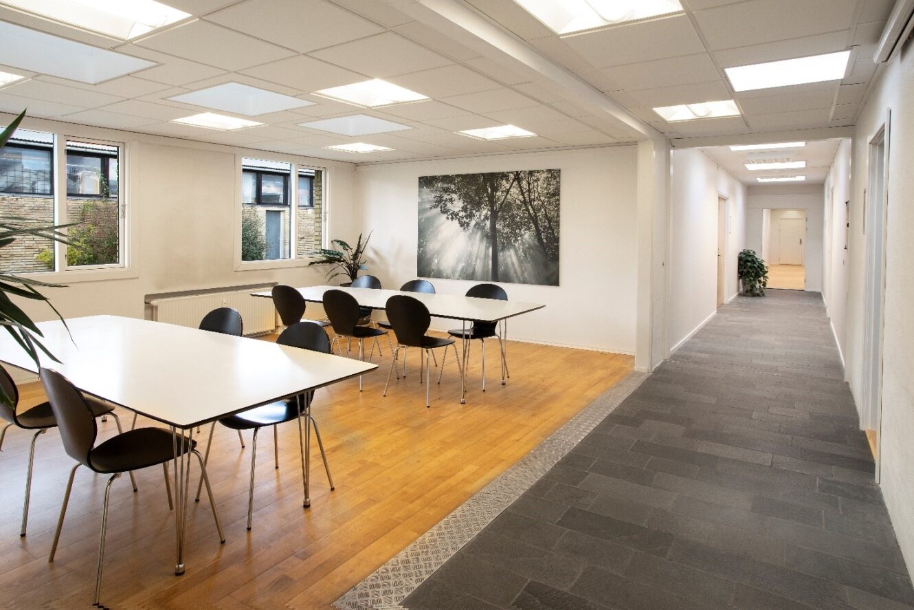 Et lyst, moderne kontor-pauserum med to borde, sorte stole, store vinduer, vægkunst og en gang, der fører til lukkede døre.