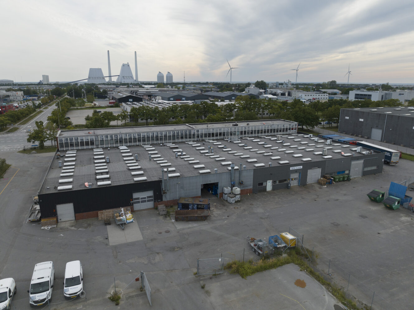 Et industrilager med fladt tag ligger på en asfalteret grund, omgivet af parkerede køretøjer og industrielt udstyr; vindmøller og kraftværksstrukturer kan ses i det fjerne.