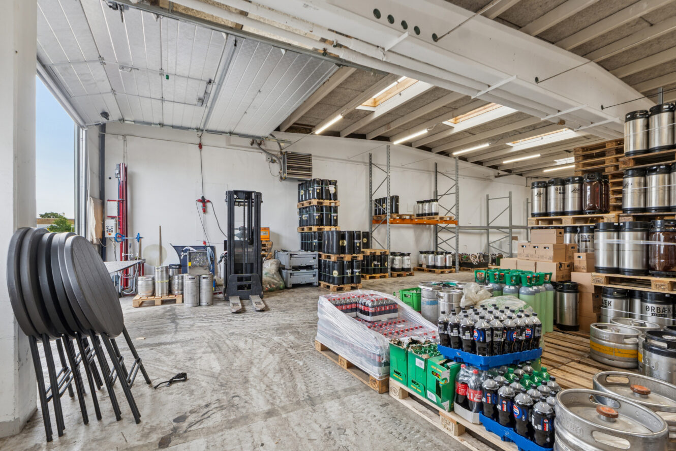 Et lagerområde med paller med drikkevarer på flaske, fustager, industrielle hylder, en gaffeltruck og stablede borde under en delvist åben garageport.