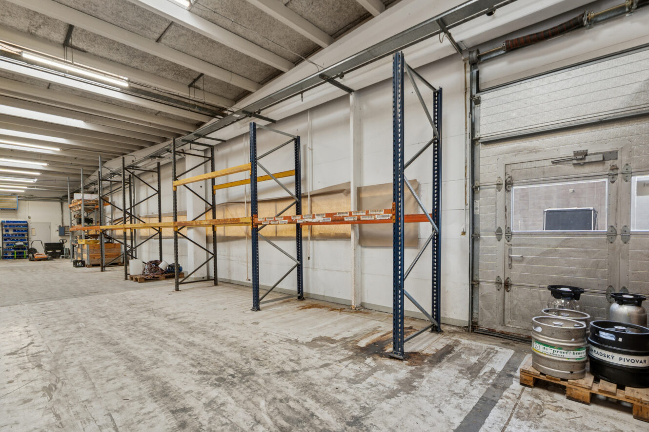 Tomme metalreoler i en rummelig lagerbygning med betongulv, højt til loftet og et par øltønder stablet på en palle nær en lukket garageport.