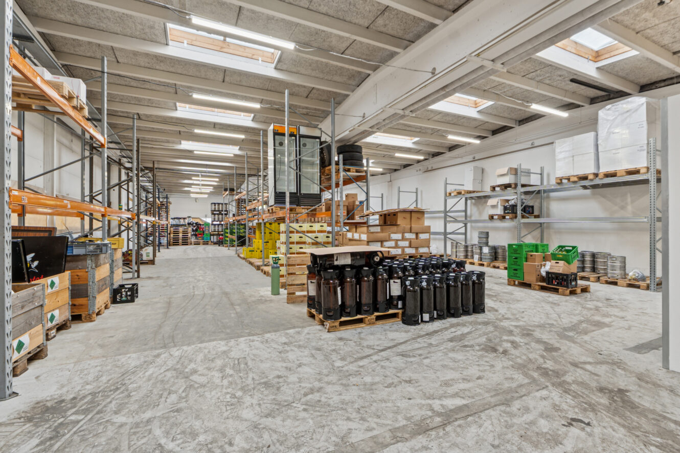 Bredt billede af et lagerinteriør med reoler, paller, lagercontainere og forskellige industrielle materialer arrangeret i hele rummet.