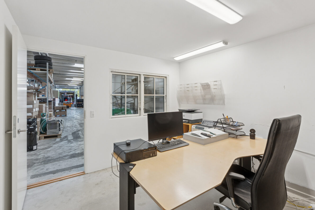 Et simpelt kontor med skrivebord, stol, computer og kontorartikler, der støder op til en lagerbygning, som man kan se gennem en åben dør og et vindue.