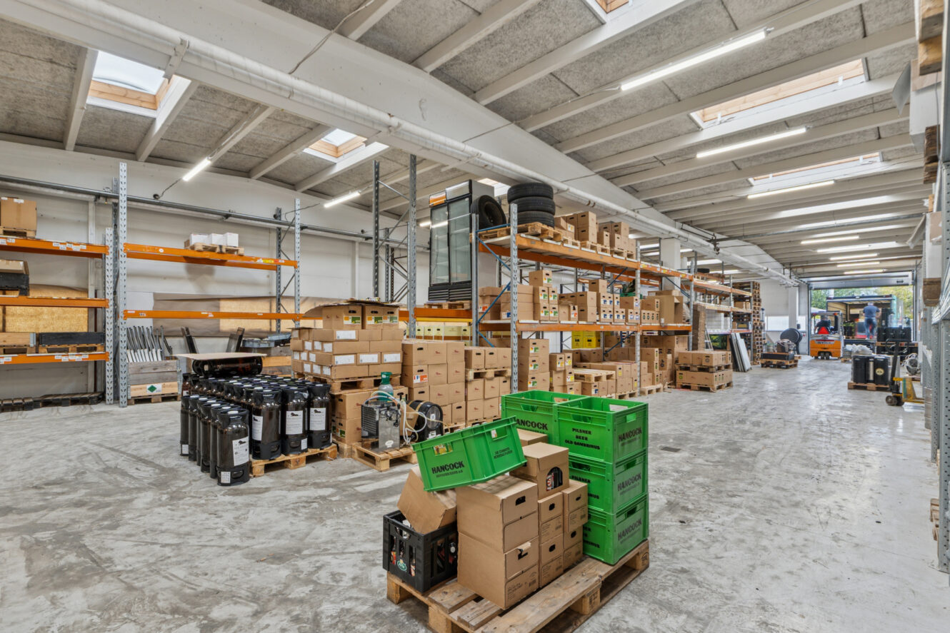 Et rummeligt lagerinteriør med hylder og paller stablet med papkasser, grønne kasser og forskelligt udstyr under fluorescerende belysning.