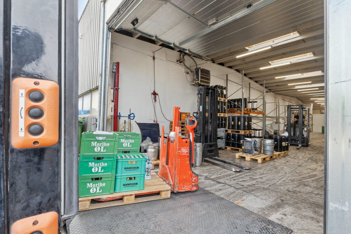 En læsserampe med grønne kasser med Marble ØL-øl, orange palleløftere og paller med fustager og tønder inde i en lagerbygning.