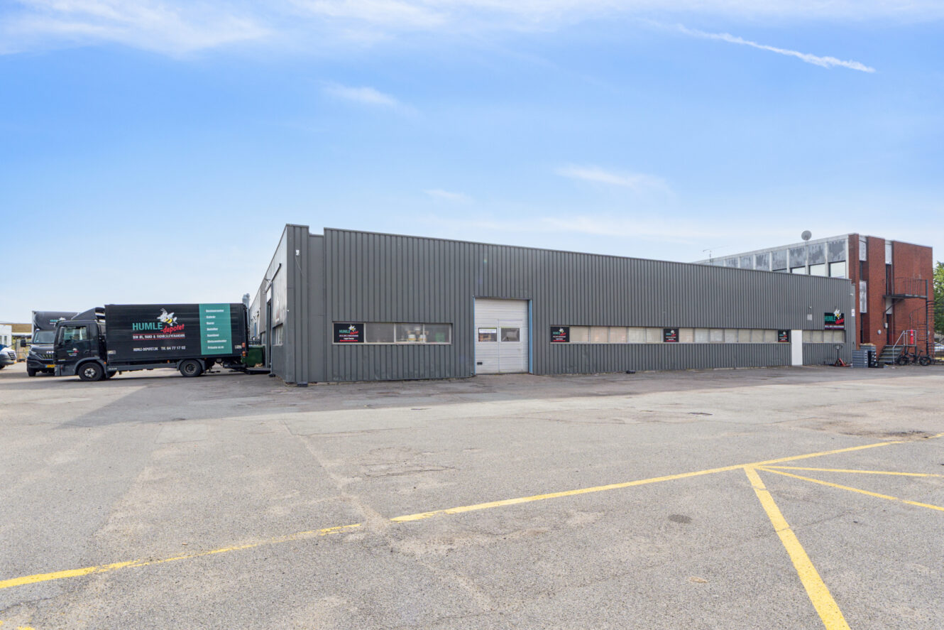 Stort, gråt industrilager med en lastbil parkeret til venstre, beliggende på en rummelig, stort set tom parkeringsplads under en klar himmel.
