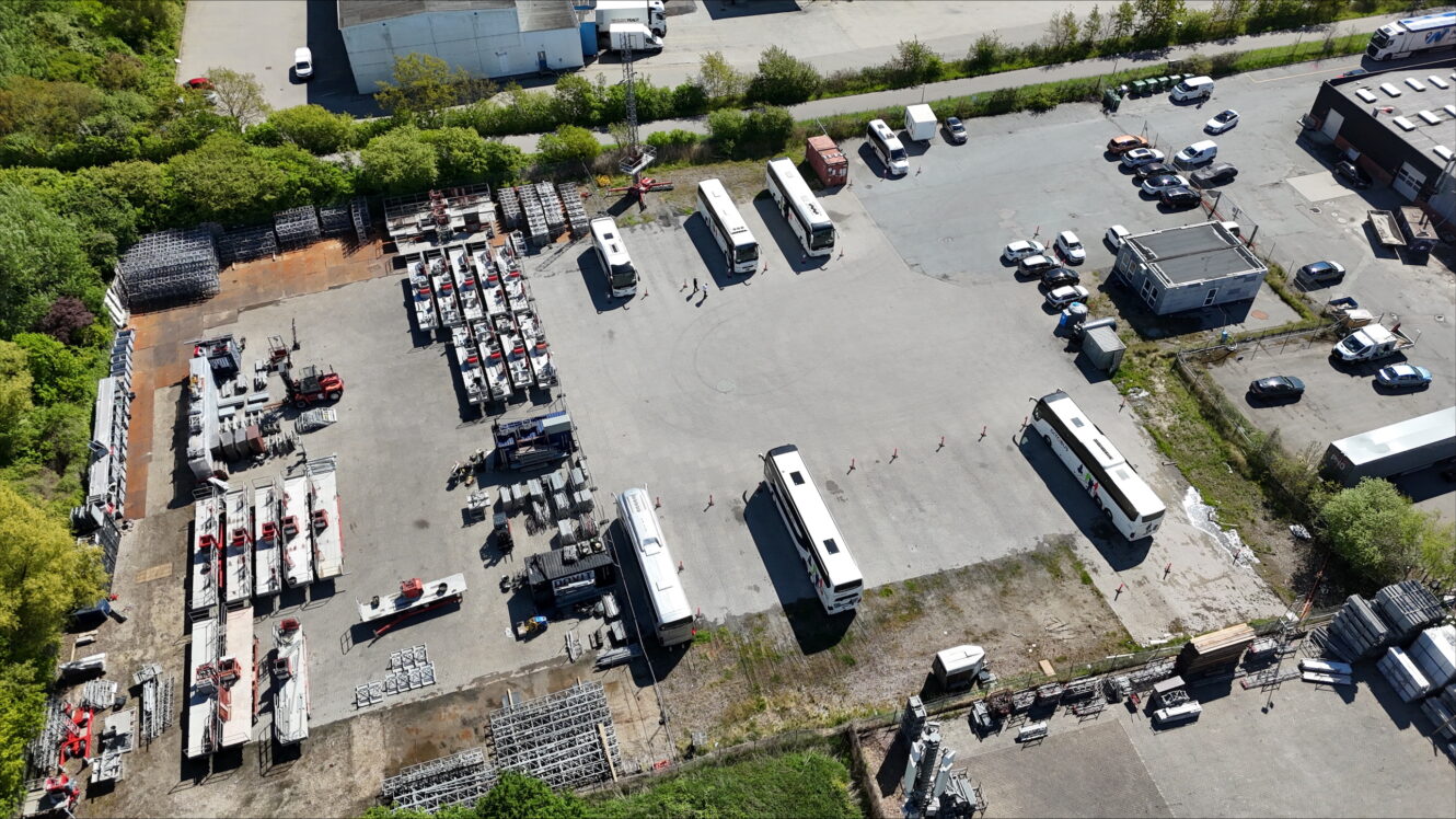 Luftfoto af en stor åben grund med flere parkerede busser, lastbiler, biler, lagerreoler, byggematerialer og små bygninger omgivet af træer og industristrukturer.