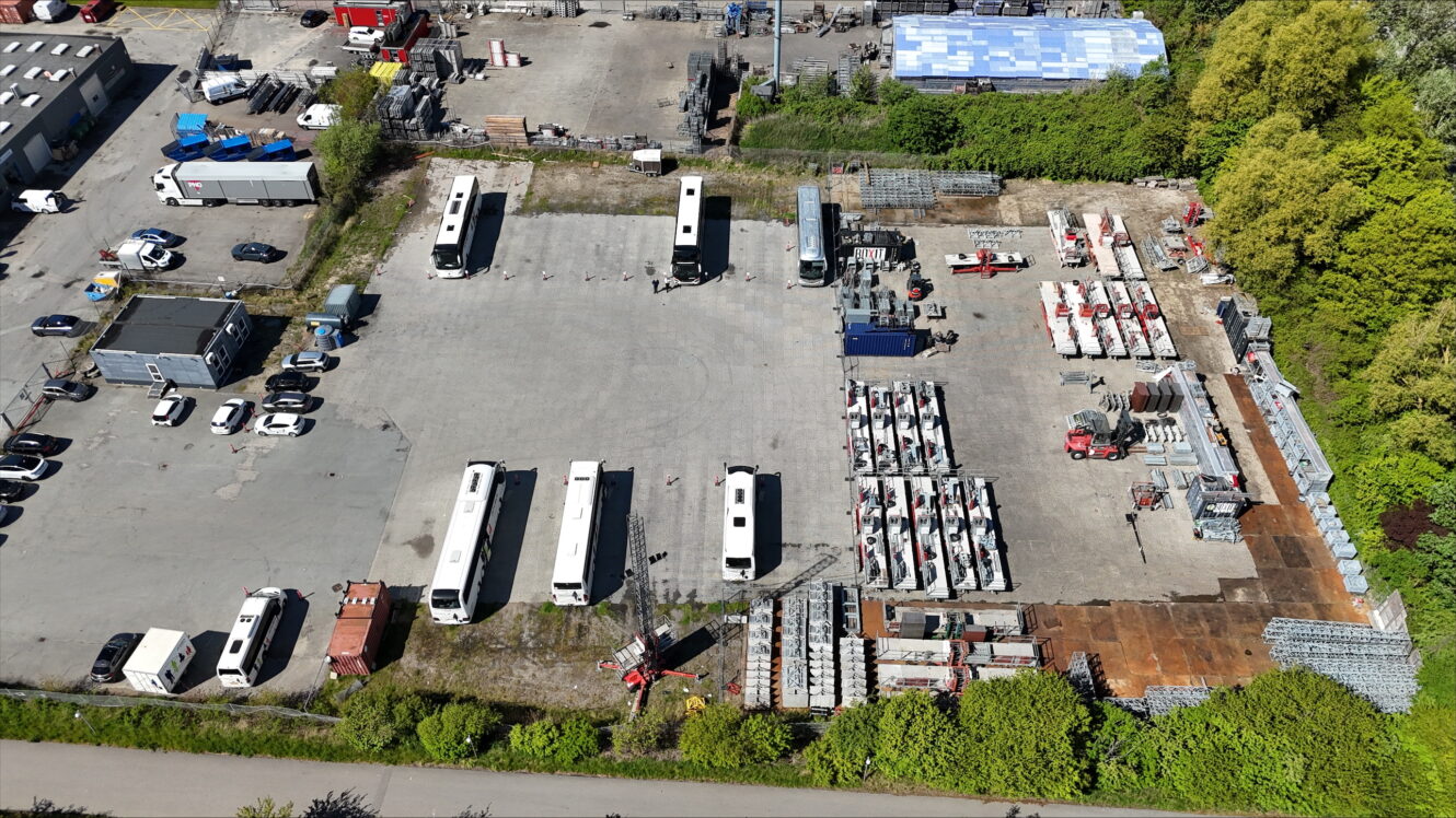 Luftfoto af en industrigrund med flere hvide busser, parkerede biler, stablede metalrør og byggeudstyr omgivet af træer og bygninger.