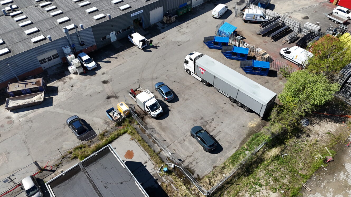 Luftfoto af en industrigård med lastbiler, biler, containere og spredt udstyr i nærheden af en lagerbygning.