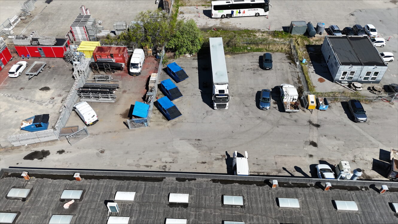 Luftfoto af en parkeringsplads med flere lastbiler, biler, blå skibscontainere, en bus, en lille bygning og spredt udstyr.