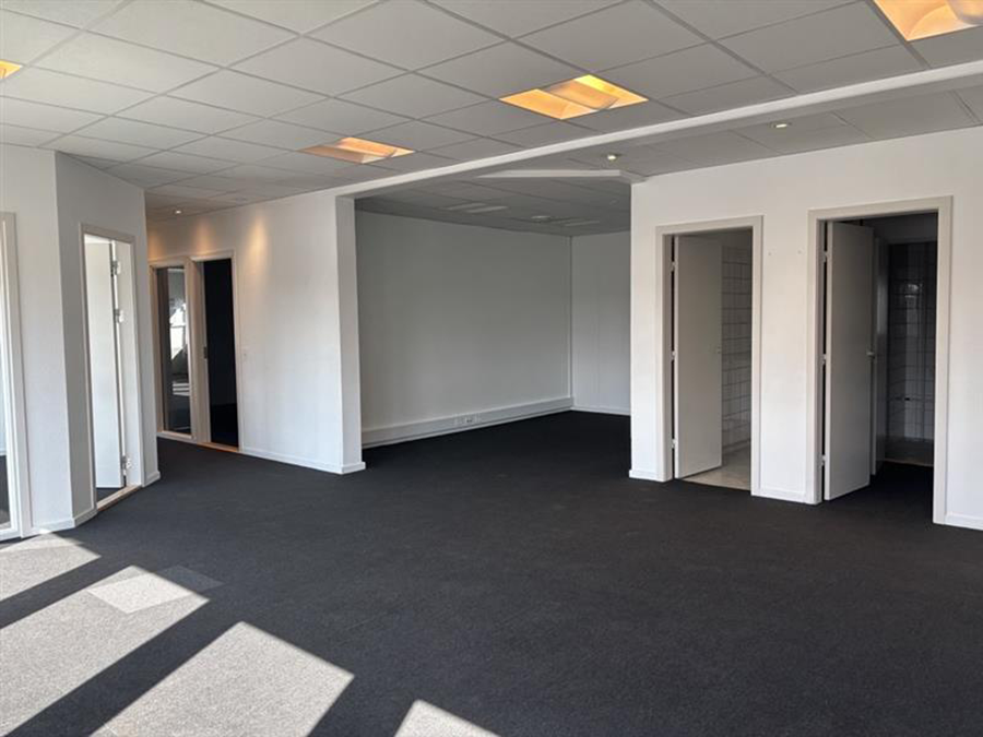 Tomt kontorlokale med gulvtæppe, hvide vægge, loftslamper, store vinduer og flere åbne døråbninger, der fører til mindre rum.