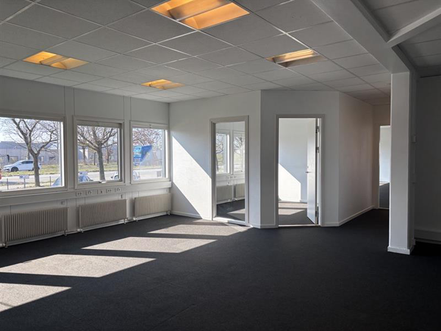Tomme kontorlokaler med store vinduer, hvide vægge, sort gulvtæppe og flere åbne døråbninger, oplyst af naturligt dagslys og loftslamper.