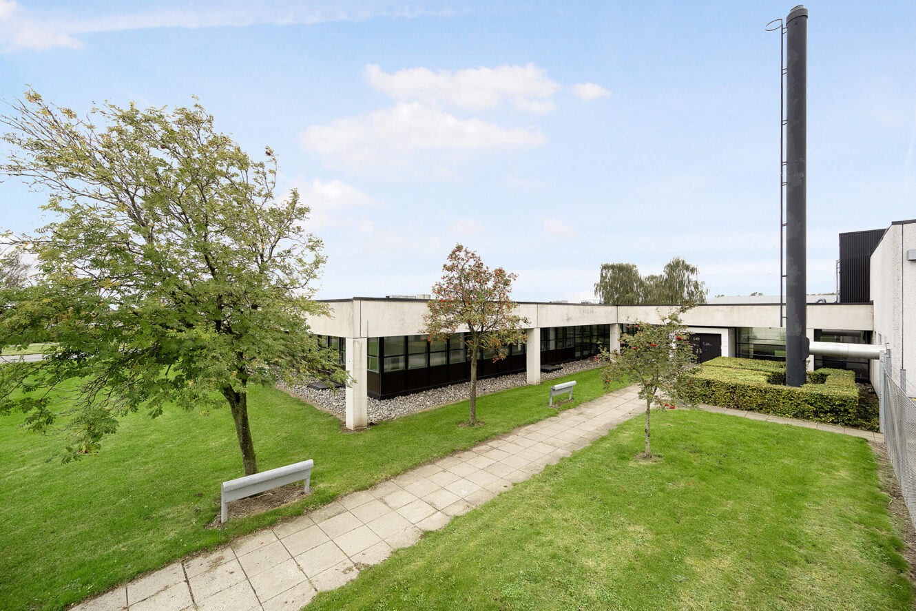 Moderne bygning i én etage med fladt tag, store vinduer og en tilstødende grøn plæne med træer, bænke og en asfalteret gangbro under en delvist overskyet himmel.