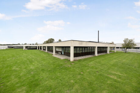 En moderne kontor- eller industribygning i én etage med store vinduer og fladt tag, omgivet af en velholdt græsplæne under en delvis overskyet himmel.