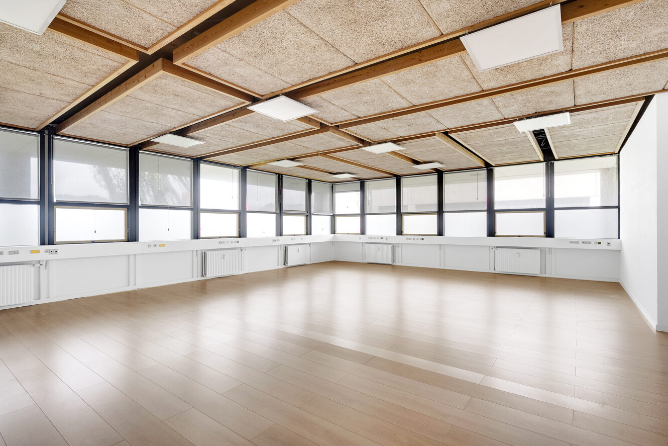 Tomt, moderne rum med store vinduer, lyse trægulve og et gittermønstret loft med indbyggede lyspaneler.