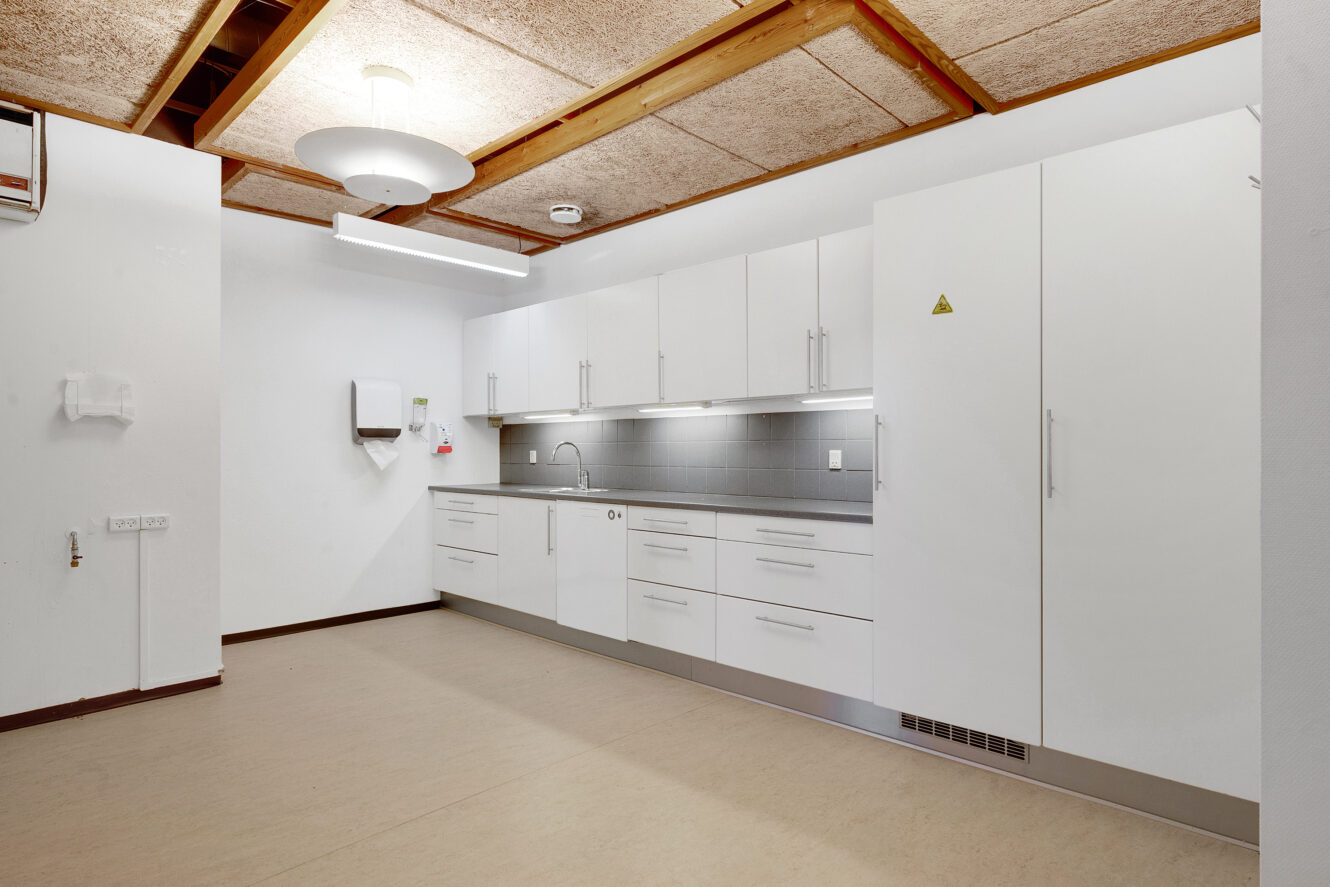 Et moderne, minimalistisk tekøkken med hvide skabe, grå backsplash, en vask og indbyggede apparater under et loft med synlige bjælker og akustiske paneler.