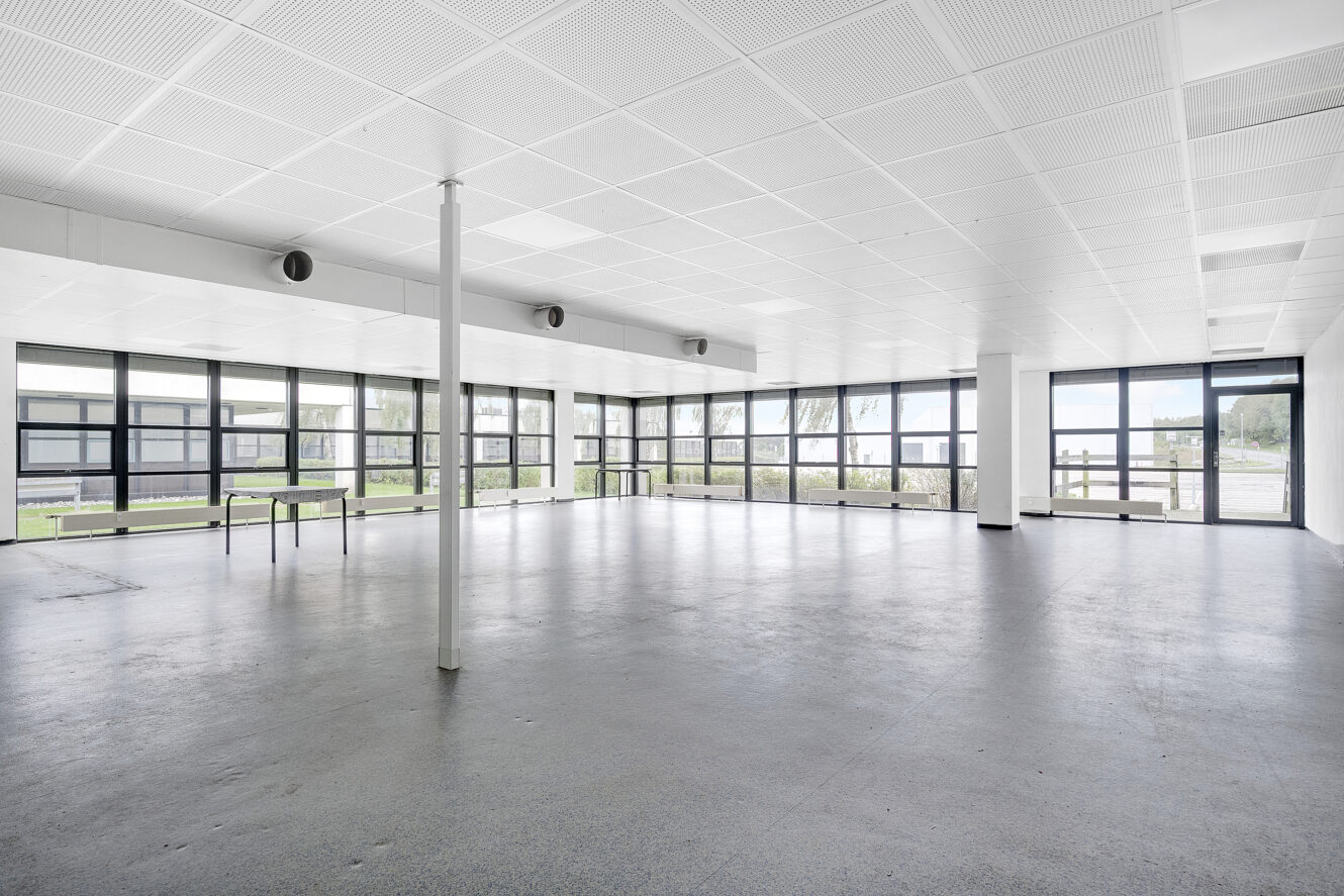 Stort, tomt, moderne rum med hvidt loft, gråt gulv, store vinduer på to sider og en enkelt søjle i forgrunden.