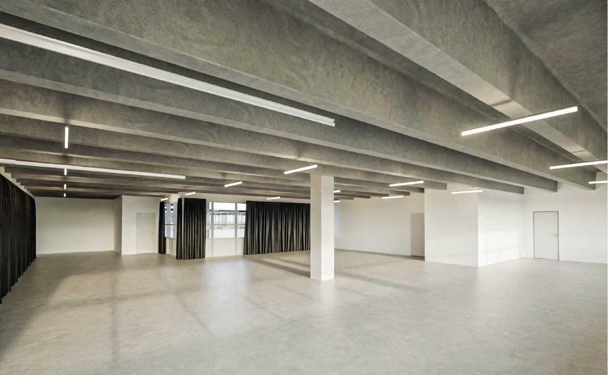 Rummeligt, tomt lokale med betongulv og synlige loftsbjælker, hvide vægge, stort vindue med mørke gardiner og lysstofrør i loftet.