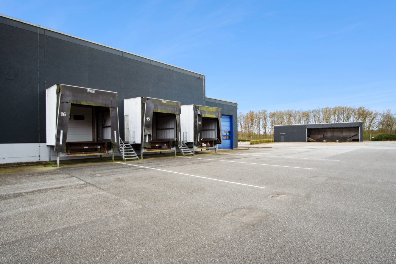 Læssedokker og ramper ved et stort industrilager med en stort set tom parkeringsplads foran under en klar blå himmel.