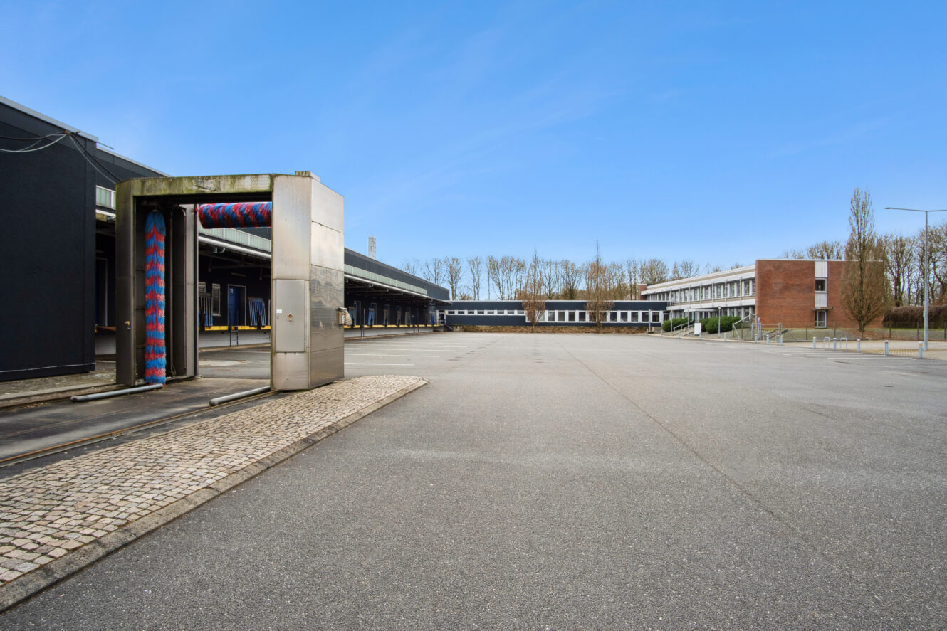 Tom industrigrund med en overdækket automatisk bilvask til venstre, lagerbygninger i baggrunden og klar blå himmel over.