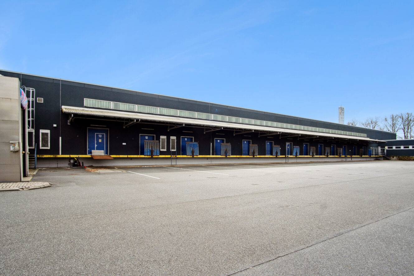 En stor industriel læsserampe med flere blå døre og hævede platforme, der støder op til en tom parkeringsplads under en klar himmel.