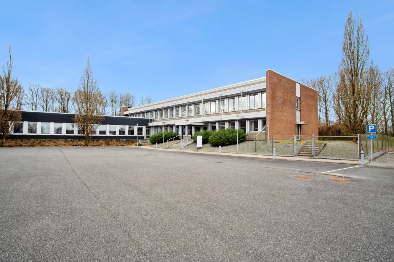 En moderne bygning i to etager med store vinduer og mursten, omgivet af nøgne træer og en tom asfalteret parkeringsplads under en klar blå himmel.