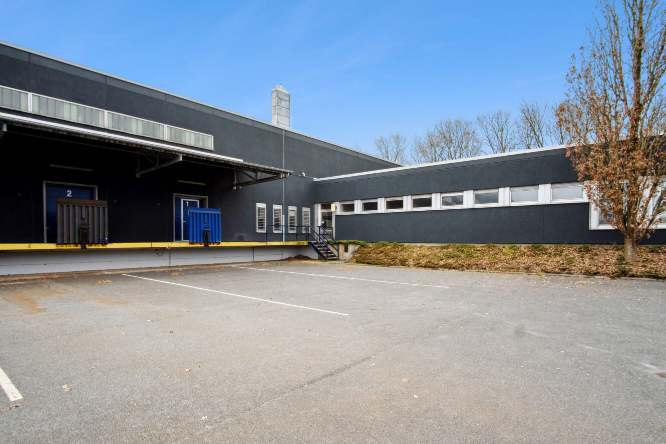 En tom parkeringsplads foran en grå industribygning med blå døre, læsseramper, trapper og bladløse træer i baggrunden under en klar himmel.