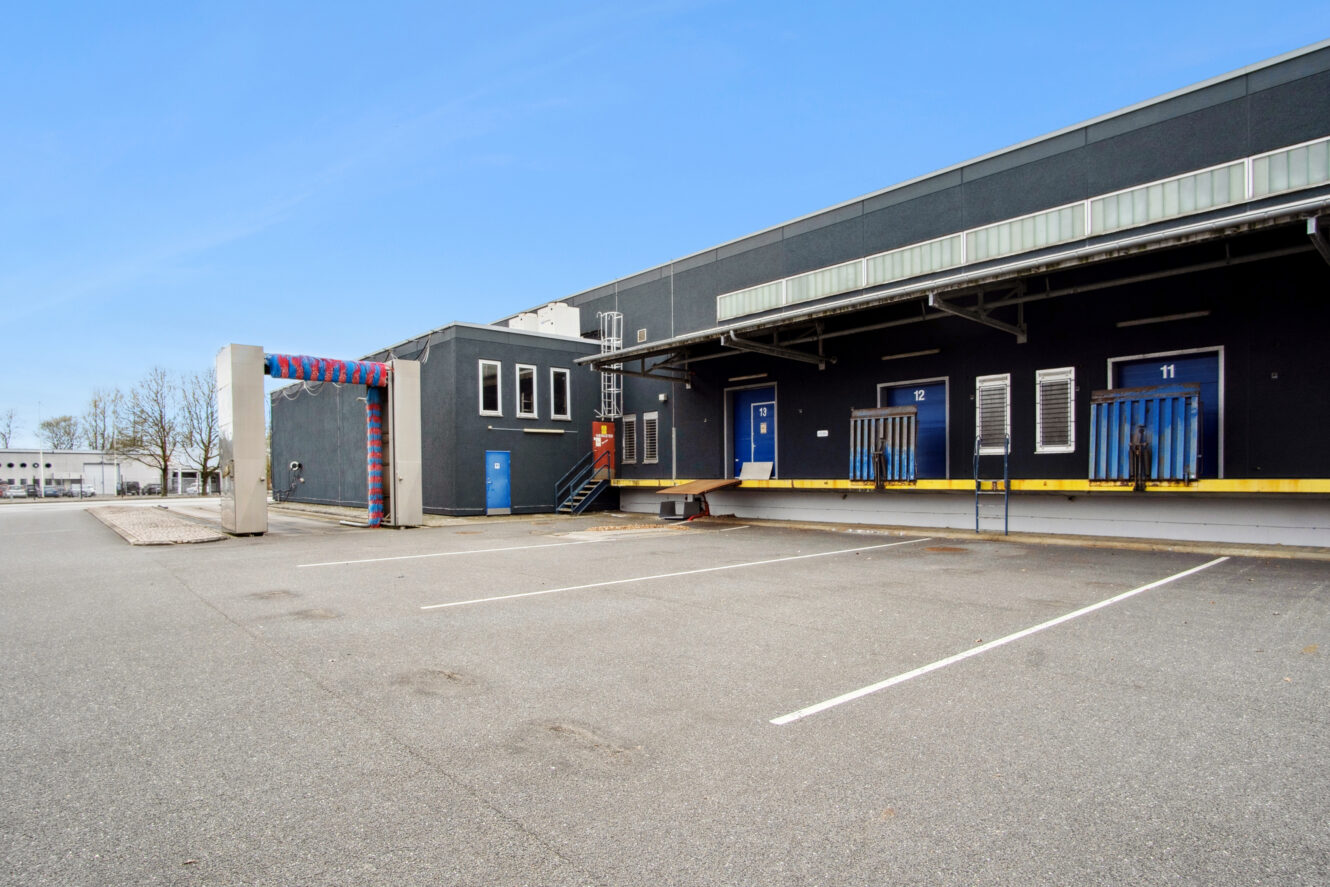 En lagerbygning med læsseramper nummer 11 og 12, en bilvaskestation til venstre og en tom parkeringsplads foran under en klar blå himmel.