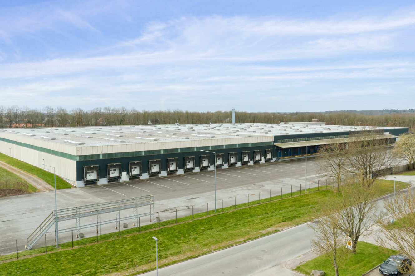Et stort industrilager med flere læsseramper og en stort set tom parkeringsplads, omgivet af træer og grønne områder under en blå himmel.