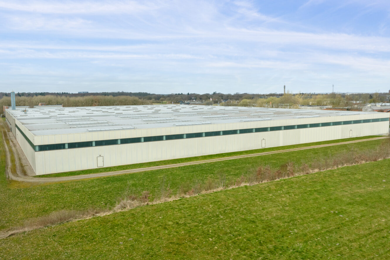 Et stort, rektangulært industrilager med fladt tag ligger på en græsklædt grund under en delvis overskyet himmel.