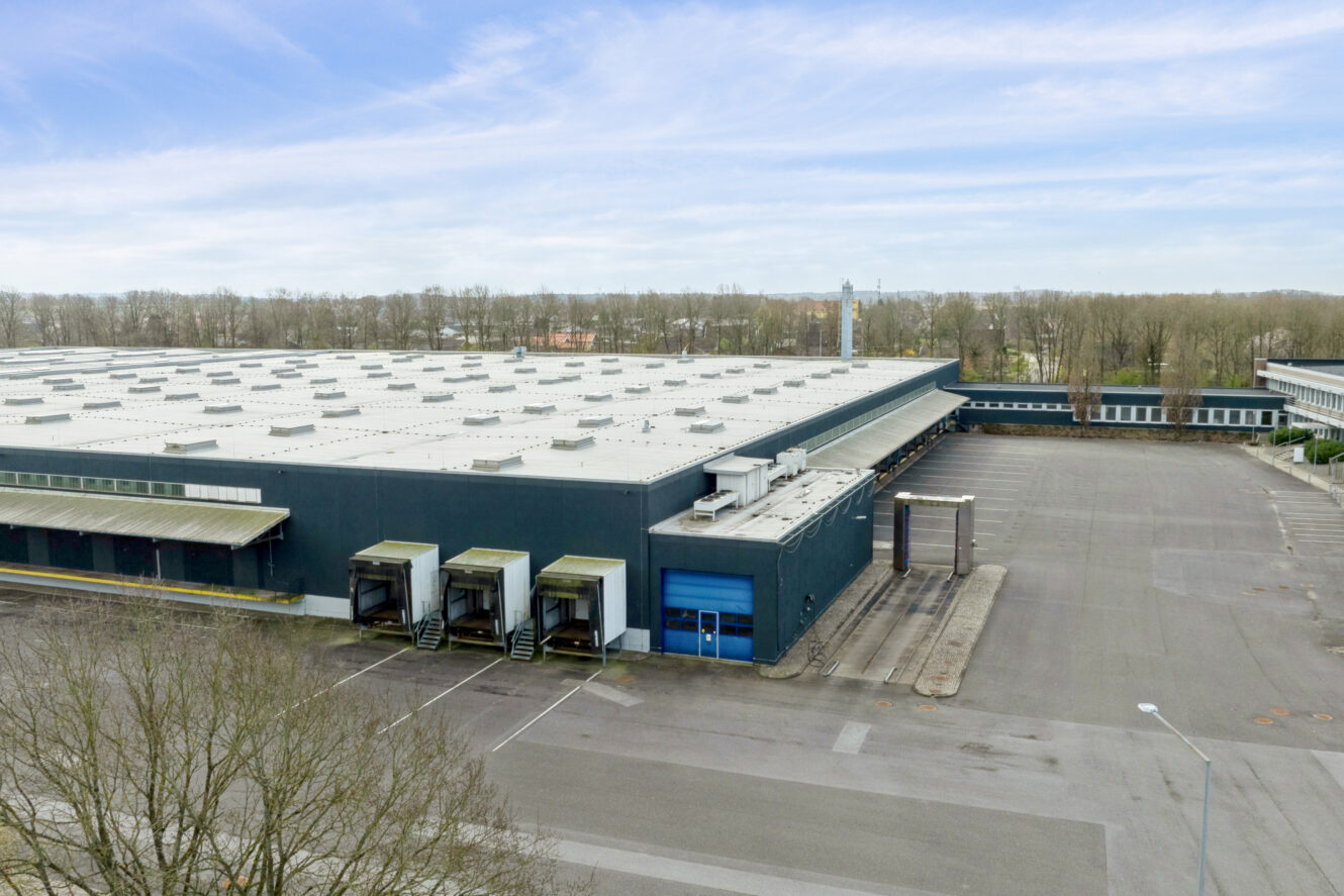 En stor, moderne lagerbygning med flere læsseramper og en tom parkeringsplads under en delvis overskyet himmel.