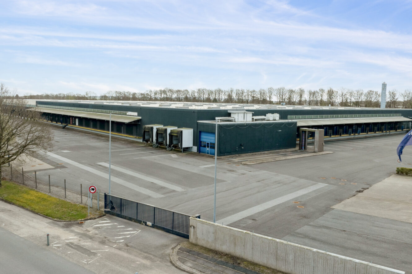 Stort industrilager med flere læsseramper, en tom parkeringsplads og et indhegnet område under en delvis overskyet himmel.