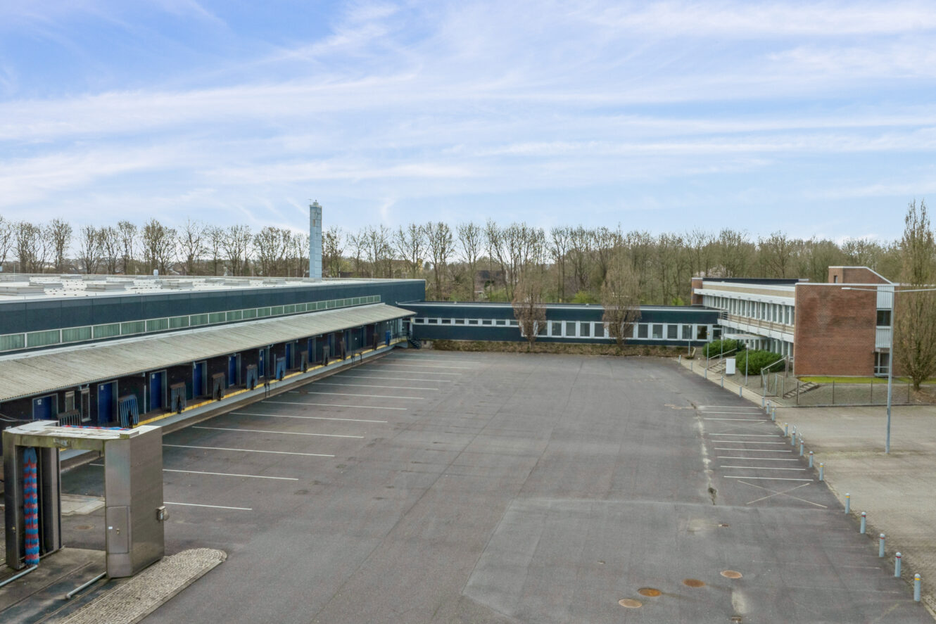 Stor, tom parkeringsplads mellem to industribygninger med træer og en overskyet himmel i baggrunden.