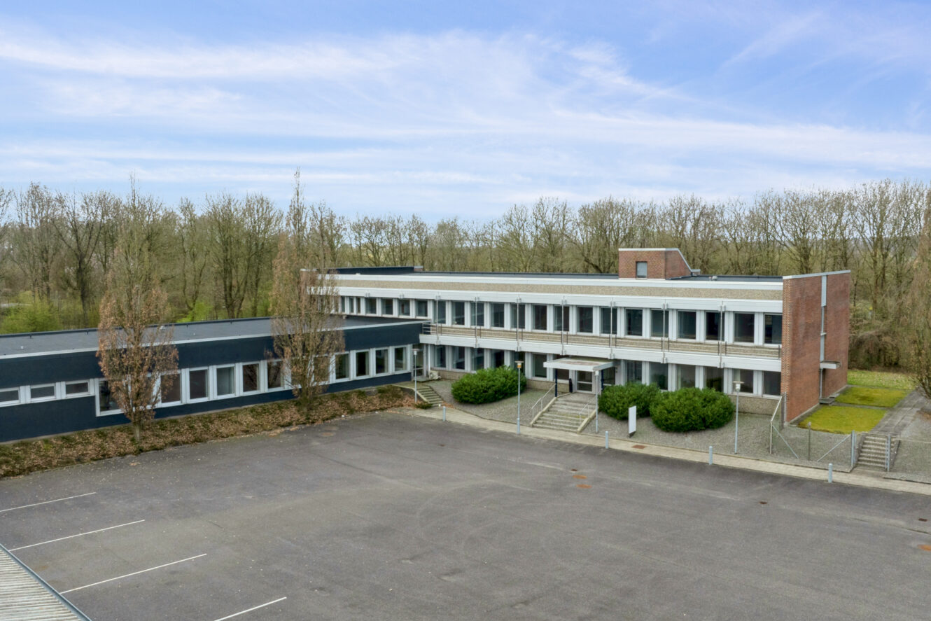 En moderne bygning i to etager med store vinduer, omgivet af træer, ligger ved siden af en tom parkeringsplads under en delvis overskyet himmel.