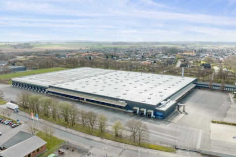 Et stort industrilager med fladt tag ligger på en stor grund, omgivet af træer og veje, med et boligområde og marker i baggrunden.