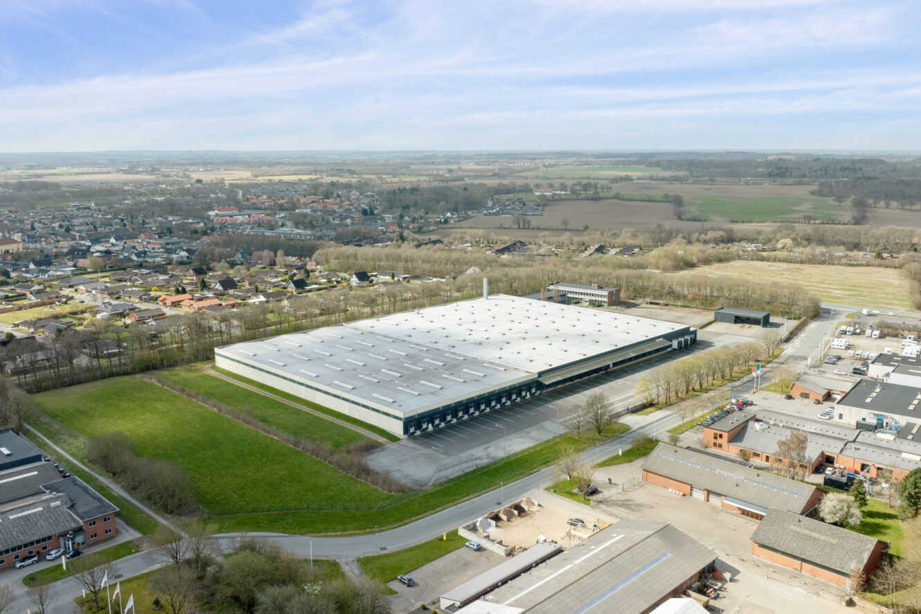 Luftfoto af et stort industrilager omgivet af grønne marker, veje og mindre bygninger i et semi-landligt område.