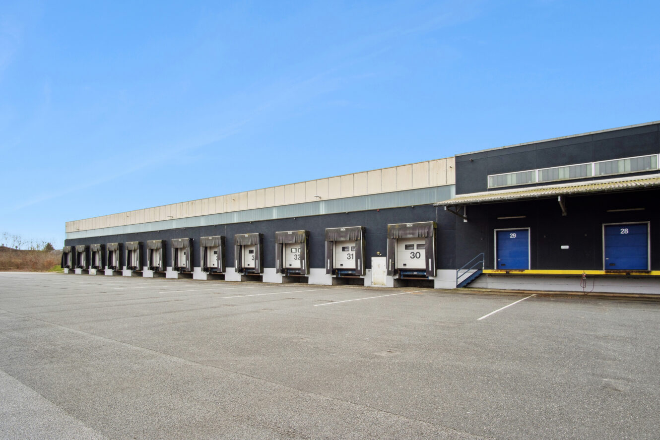 En stor industribygning med flere læsseramper og garageporte, der støder op til en tom parkeringsplads under en klar blå himmel.