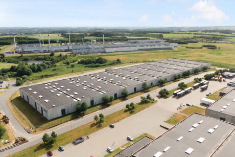 Luftfoto af store industrilagre omgivet af veje, lastbiler og grønne marker med flere bygninger og vindmøller i baggrunden.