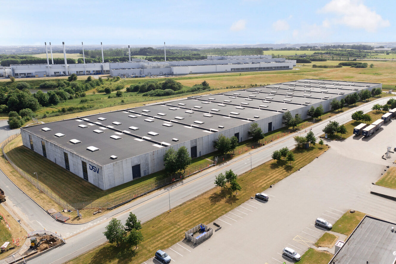 Luftfoto af et stort industrilager med fladt tag, omgivet af veje, parkeringspladser og græsmarker, med en anden fabrik i baggrunden.