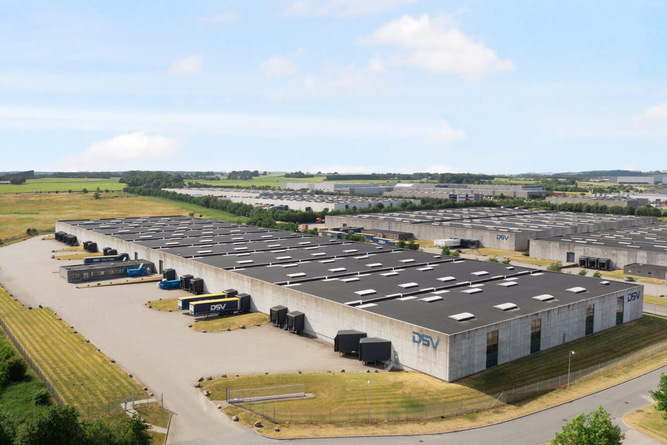 Et stort industrilager med fladt tag, flere læsseramper og flere lastbiler parkeret udenfor, omgivet af åbent land og yderligere lagerbygninger i baggrunden.